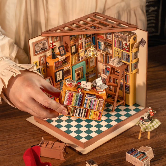 Rolife Sam's Study Library DG102 DIY miniature house shot from above with a hand placing a rainbow-colored book table against the checkered green tile floor